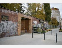 Ziegelsteinmauer mit Tor und Graffiti, daneben ein Blumenverkaufsstand mit Blumen und Pflanzen. Im Hintergrund herbstliche Bäume und ein mehrstöckiges Gebäude mit verblasster Wandbemalung.