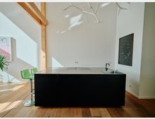 Moderne Küche mit einer schwarzen Kücheninsel, die ein Spülbecken und eine helle Arbeitsfläche integriert. Der Raum hat hohe, weiße Wände, sichtbare Holzbalken, einen Holzboden sowie dekorative Elemente wie grüne Barhocker und hängende Beleuchtung in organischem Design.