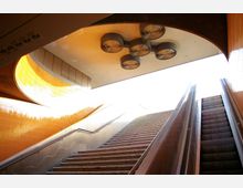 Treppenaufgang und Rolltreppenaufgang in einer U-Bahn-Station mit orangefarbenen, gefliesten Wänden und modernen, runden Deckenleuchten. Helles Tageslicht fällt durch die offene Oberseite des Übergangs ein.