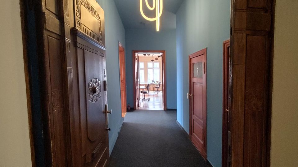 Langer Flur in einem Altbau mit blau gestrichenen Wänden, dunklem Teppichboden und großen, dekorativ geschnitzten Holztüren. An der Decke hängt eine moderne Leuchte, am Ende führt eine offene Tür in einen hellen Raum mit großen Fenstern.