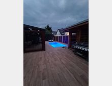Ein moderner Außenbereich mit Pool, umgeben von Holzdielen und Holzhütten. Im Hintergrund sind Wohnhäuser und eine Hecke zu sehen, und auf der rechten Seite befindet sich ein Grill.