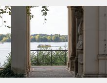 Blick durch einen Säulengang auf einen Fluss mit einem gelben Ausflugsboot und einer bewaldeten Uferlandschaft im Hintergrund. Im Vordergrund ist ein Geländer mit Ornamenten sowie ein gemusterter Steinboden zu sehen.