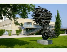 Moderne Skulptur auf einer Grünfläche vor einem Gebäude mit heller Fassade und großen Fenstern; links eine Baumkrone, rechts ein schlanker Baum. Im Hintergrund führt eine Außentreppe zum oberen Stockwerk des Gebäudes, über dessen Eingang ein Schild mit der Aufschrift "Haveldeck" angebracht ist.