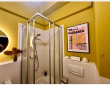 Ein Badezimmer mit gelb gestrichenen Wänden und weiß gefliesten Bereichen. Im Vordergrund befindet sich eine Duschkabine mit Glaswänden, daneben ein beleuchteter runder Spiegel, eine Vase mit Trockenblumen und ein Kunstposter von Yayoi Kusama an der Wand.