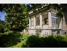 Seitenansicht einer älteren Villa mit überdachter Veranda, getragen von mehreren dicken Säulen, und großen Sprossenfenstern. Davor liegt ein üppiger Garten mit Stauden und hohen Bäumen, neben einem schmalen Weg.