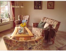 Wohnzimmer mit gemütlicher Einrichtung, bestehend aus einem Holztisch und einer Sitzgruppe mit floral gemusterten Polstermöbeln. Ein großes Fenster mit Vorhängen lässt Licht herein, und auf der Fensterbank stehen Pflanzen; an der Wand hängen zwei Bilder.