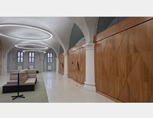 Moderner Innenraum einer Lobby oder Wartehalle mit hohen Gewölbedecken, weißen Säulen und großen ringförmigen Deckenleuchten. Rechts verläuft eine lange Holzvertäfelung mit geometrischem Muster, links stehen Sitzbänke und kleine Tische vor hohen Fenstern.