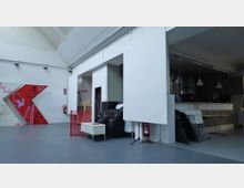 Großer, moderner Raum mit grauem Boden, weiß gestrichenen Wänden und hohen Decken. Links befindet sich eine bemalte Wand mit roten und rosa Grafiken, rechts eine Theke mit Hängelampen, dahinter Stühle und Tische gestapelt an der Wand.