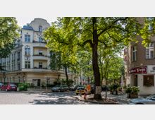 Kreuzung einer ruhigen, mit Kopfsteinpflaster ausgelegten Straße in einer Wohngegend, umgeben von historischen mehrstöckigen Gebäuden mit Balkonen und verzierten Fassaden. Im Vordergrund stehen große, schattenspendende Bäume, und rechts ist eine Gaststätte mit dem Schriftzug "Zur alten Post" zu sehen.