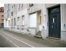 Eine graue Wohnhausfassade in einer urbanen Umgebung mit mehreren Fenstern und zwei Eingangstüren, von denen eine schwarz und die andere weiß ist. Die Wände sind teilweise mit Graffiti bedeckt, und der Gehweg davor besteht aus Pflastersteinen und einem Radweg.