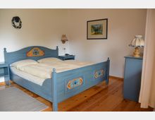 Schlafzimmer mit Holzboden und einem blauen Doppelbett im Landhausstil, verziert mit floralen Mustermalereien. Im Raum befinden sich passende Nachttische, eine Wandlampe, ein gerahmtes Landschaftsbild sowie Dekoration an den Wänden.