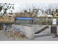 Eingang zur U-Bahn-Station Mierendorffplatz mit einem blauen Schild, das den Stationsnamen zeigt. Im Vordergrund stehen mehrere abgestellte Fahrräder, umgeben von Bäumen mit kahlen Ästen und herabgefallenen Blättern.