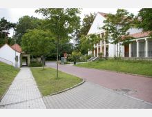 Weißes, zweistöckiges Gebäude mit Säulenvorbau und roten Ziegeldächern, umgeben von Bäumen und Grünflächen. Im Vordergrund führen gepflasterte Wege und ein kleiner Zebrastreifen zu dem Gebäude, während ein „Stop“-Verkehrsschild nahe der Einfahrt steht.