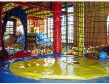 Ein Indoor-Spielplatz mit bunten Bällen in einem Bällebad, umgeben von Netzen und großen, farbenfrohen Kletterstrukturen aus Kunststoff. Im Vordergrund befindet sich eine flache Sprungfläche.