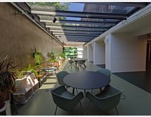 Langgestreckter Innenraum mit Glasdach und modernem Design; links eine Betonwand mit zahlreichen Topfpflanzen auf Regalen, rechts weiße Wände mit offenen Durchgängen. Im Vordergrund und entlang des Raumes stehen Tische mit Stühlen, im Hintergrund eine grün gemusterte Wand.