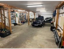 Eine helle Werkstatt oder Garage mit einem alten schwarzen Cabriolet in der Mitte. Holzstützen, Regale mit Werkzeugen und Materialien, sowie Werkbänke und diverse Gegenstände wie Rohre und Möbelstücke prägen den Raum.