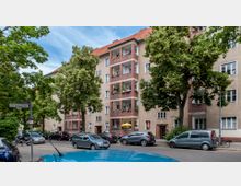 Wohnhaus in einer urbanen Straße mit drei Stockwerken und Balkonen, die mit Pflanzen und Dekorationen geschmückt sind. Umgeben von Bäumen und geparkten Autos, mit einem Straßenschild der „Gritznerstraße“ im Vordergrund.