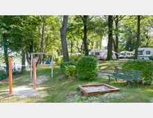 Campingplatz in einer bewaldeten Umgebung mit Wohnmobilen und Zelten im Hintergrund. Im Vordergrund befinden sich ein Spielplatzbereich mit einer Reckstange, ein Sandkasten und eine grüne Bank, umgeben von Sträuchern und hohen Bäumen.