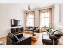 Wohnzimmer mit moderner Einrichtung, darunter schwarze Ledersessel und ein Sofa, ein hölzerner Couchtisch und ein dunkler TV-Schrank mit einem wandmontierten Flachbildfernseher. Große Fenster mit beigefarbenen Vorhängen lassen Tageslicht einfallen, und eine Leuchte mit drei hängenden Glaslampen hängt von der Decke.
