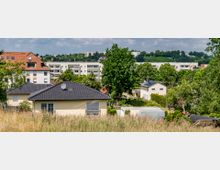 Wohngebiet mit Einfamilienhäusern im Vordergrund, umgeben von Gärten und Bäumen. Im Hintergrund sind mehrstöckige Wohngebäude, eine hügelige Landschaft und vereinzelt Weinberge zu sehen.