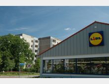 Ein Lidl-Supermarkt mit grauer Fassade und einem roten Ziegeldach, im Vordergrund Schaufenster mit Werbepostern. Im Hintergrund stehen mehrstöckige Wohnblöcke und Bäume, unter einem blauen Himmel.
