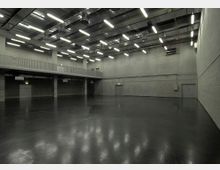 Großer, leerer Innenraum mit Betonwänden, schwarzem glänzendem Boden und industrieller Decke, die mit sichtbaren Lüftungsrohren und langen, rechteckigen Leuchtstofflampen ausgestattet ist. Auf der linken Seite befindet sich eine erhöhte Galerie mit Metallgeländer.