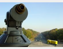 Ein silbernes Fernglas auf einer Plattform im Vordergrund, blickend auf eine lange, baumgesäumte Straße in Berlin mit Autos und einem gelben LKW; im Hintergrund ist der Berliner Fernsehturm sichtbar.