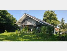 Modernes Einfamilienhaus mit Satteldach und großen Fensterfronten, umgeben von einem gepflegten Garten mit Bäumen, Sträuchern und Rasenfläche. Im Hintergrund befinden sich weitere Vegetation und vereinzelte Gebäude.