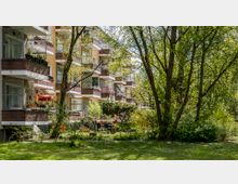 Wohngebäude mit mehreren Balkonen, die mit Pflanzen und Dekorationen geschmückt sind, umgeben von einer Grünanlage mit Bäumen, Sträuchern und einem kleinen Sitzbereich im Garten. Die Szenerie zeigt eine ruhige, naturreiche Umgebung.