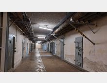 Ein langer, schmaler Korridor mit abblätternden Wänden, schweren Metalltüren und freiliegenden Rohrleitungen und Kabeln an der Decke. Die Beleuchtung erfolgt durch Neonlampen, und der Boden wirkt abgenutzt und feucht.