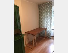 Ein kleiner Raum mit hellem Laminatboden, einer weißen Wand, einem braunen Holztisch mit einfacher Konstruktion und einem Vorhang mit Blattmuster vor einem Fenster. Links steht ein grünes Holzregal.