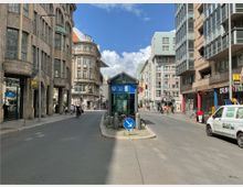 Straßenszene in einer Stadt mit mehrstöckigen Gebäuden im historischen und modernen Stil. Im Vordergrund befindet sich ein gläserner U-Bahn-Eingang mit einem blauen Schild; im Hintergrund sind Passanten, Radfahrer und ein Schild mit der Aufschrift „Haus am Checkpoint Charlie“ erkennbar.