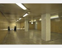 Unterirdischer Fußgängertunnel mit gefliesten Wänden in Beige- und Weißtönen sowie rechteckigen weißen Säulen. Der Boden besteht aus quadratischen Steinfliesen, und Neonlichter an der Decke beleuchten den Raum; zwei Rolltreppen führen nach unten.