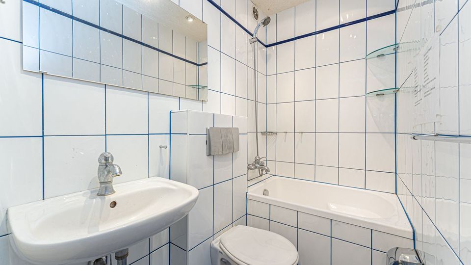 Kleines, helles Badezimmer mit weißen Wandfliesen und blauen Fugen, ausgestattet mit Waschbecken, WC und Badewanne mit Duscharmatur. An der Wand befinden sich ein großer Spiegel sowie Glasablagen.