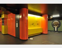 Untergrundstation mit bunten Wänden in Gelb, Orange und Schwarz, beleuchtet von Deckenleuchten. Eine Sitzbank ist in eine gelbe Wand integriert, die mit Graffiti bedeckt ist, und eine runde Wanddekoration zeigt ein gemaltes historisches Stadtmotiv.