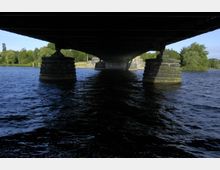 Blick unter eine Brücke über einen Fluss mit massiven, steinernen Pfeilern. Im Hintergrund sind Bäume und Uferbereiche erkennbar, die bei Tageslicht beleuchtet sind.