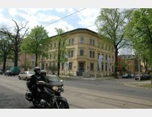 Eckgebäude im Stil des späten 19. Jahrhunderts mit gelber Fassade und hohen Fenstern, umgeben von Bäumen. Im Vordergrund eine Straße mit Straßenbahnschienen, vorbeifahrende Fahrzeuge und ein Motorrad mit zwei Personen.