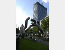 Skulptur auf einer grünen Rasenfläche in einer städtischen Umgebung, dargestellt durch ineinander greifende Metallbögen; im Hintergrund ein modernes Hochhaus mit Glasfassade und einige Bäume am Straßenrand.