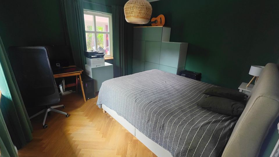 Schlafzimmer mit dunkelgrünen Wänden, Parkettboden und einem Doppelbett mit grauer Tagesdecke. Neben einem Fenster mit Vorhängen stehen ein Schreibtisch mit Computer und ein Drucker; gegenüber befindet sich ein hoher Schrank und eine geflochtene Pendelleuchte an der Decke.