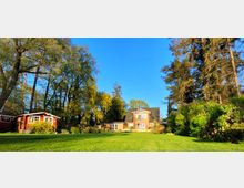 Ein Backsteinhaus mit dunkel gedecktem Satteldach inmitten eines großen Gartens mit gepflegtem Rasen, umgeben von hohen Bäumen und Büschen. Links ein kleines rotes Gartenhäuschen, ergänzt durch weitere Bepflanzung.