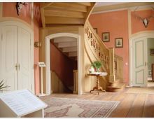 Heller Innenraum mit einer Holztreppe, die nach oben führt, rosa gestrichenen Wänden und weißen Türen mit verzierten Details. Ein kleiner Tisch mit Dekoration steht auf einem Holzboden, der mit einem orientalischen Teppich bedeckt ist, während Wandschmuck und ein Geweih ergänzt.