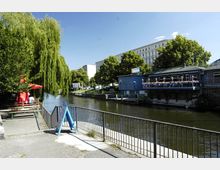 Ein Kanal mit angrenzenden Grünflächen, einer großen Trauerweide und einem Uferrestaurant mit blauer Fassade und Sitzbereich auf einer Terrasse. Im Hintergrund sind Bäume und ein mehrstöckiges Gebäude sichtbar, während der Vordergrund einen Gehweg mit Geländer und einem Wegweiser zeigt.