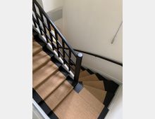 Eine Wendeltreppe mit schwarzen Stufen, die mit einem beigefarbenen Teppichläufer bedeckt sind, und einem schwarzen Geländer mit Gittermuster. Die Wände sind weiß gestrichen, und eine Beleuchtungskabelinstallation ist sichtbar.