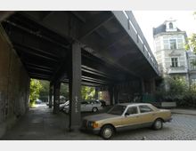 Eine stählerne Eisenbahnüberführung, die über einen schattigen Gehweg mit parkenden Autos führt. Im Hintergrund sind ein Altbau mit ornamentalen Fassaden und grüne Vegetation sichtbar.
