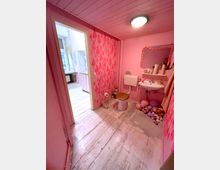 Kleines Badezimmer mit rosa Wänden, weißem Holzboden und einer Toilettenschüssel mit goldener Sitzfläche. An der Wand sind dekorative Muster und am Boden liegen bunte Luftballons; ein Waschbecken mit Spiegel und Dekorationsartikeln befindet sich rechts.