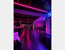 Innenraum eines modernen Club- oder Barbereichs mit dunklem Boden, langer Theke links und Sitzgruppen im Hintergrund, beleuchtet von pinken LED-Lichtleisten. Sichtbare Deckenpaneele, Stützen und hohe Stehtische entlang der rechten Wand prägen den Raum.