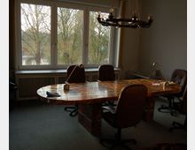 Konferenzraum mit einem ovalen Tisch aus poliertem Holz, umgeben von drehbaren Bürostühlen. Große Fenster im Hintergrund bieten den Blick auf eine baumreiche Außenszenerie und lassen Tageslicht herein; eine schwarze Kronleuchter-Lampe hängt von der Decke.