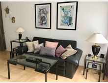 Wohnzimmer mit schwarzem Ledersofa und vielen Kissen, davor ein Glascouchtisch auf hellem Holzfußboden. An der weißen Wand hängen zwei gerahmte abstrakte Bilder; rechts und links stehen Beistelltische mit Lampen, daneben sind dekorative Masken an der Wand angebracht.