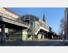 Überdachte, historische Hochbahnstation mit bogenförmigen Glasfenstern, darunter grüne Wandfliesen. Im Hintergrund ist eine Kirchturmspitze sichtbar, umgeben von Stadtarchitektur und einem klaren blauen Himmel.