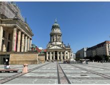 Weitläufiger Platz mit gepflastertem Boden, im Zentrum das Deutsche Domgebäude mit Kuppel, flankiert von historischen Gebäuden, darunter links das Konzerthaus Berlin mit Säulenfassade und Reiterstatuen. Klare, sonnige Atmosphäre und umgebende Bebauung im Hintergrund.